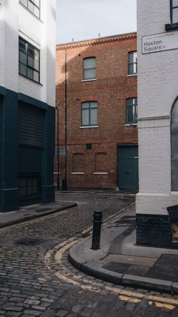 A narrow, cobblestone street lined with multi-story buildings featuring a mix of brick and white facades, with some buildings adorned with green climbing plants and window boxes. The scene is illumina